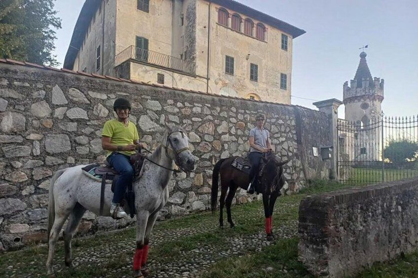 Two horses with riders, a stone wall, and a building with a tower in the background. 1