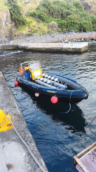 A black inflatable boat named 'AGARRÓ' docked at a concrete pier with a rocky shoreline in the background. 1