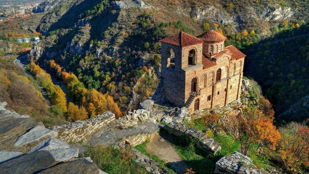 A stone church perched on a cliffside, surrounded by autumn foliage and rocky terrain. 1