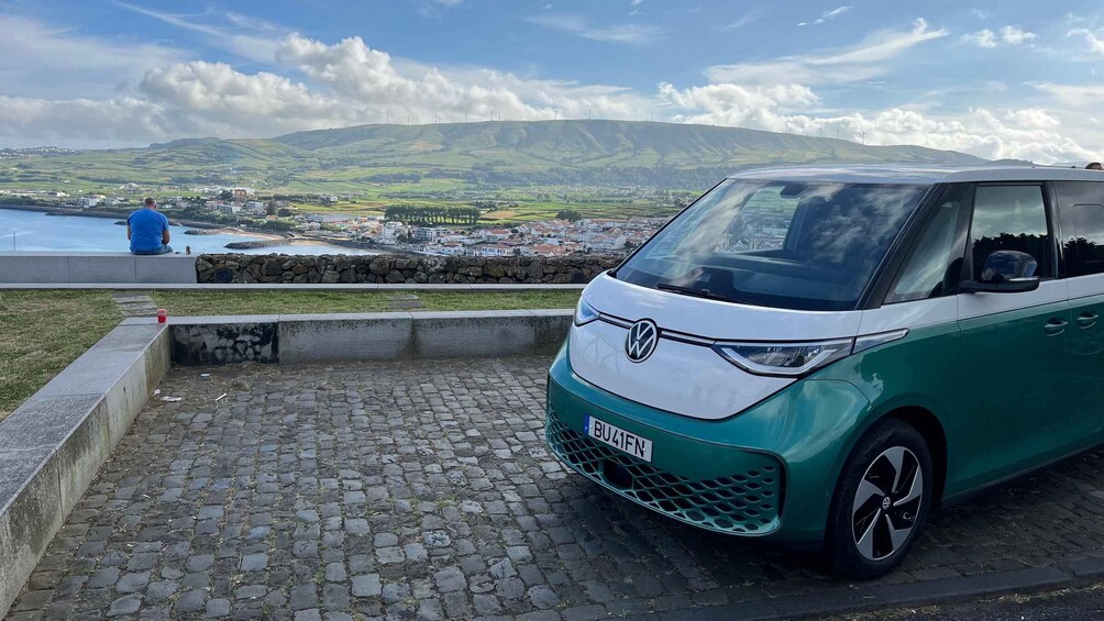A green and white car parked on a cobblestone path with a scenic view of a town and hills in the background. 1