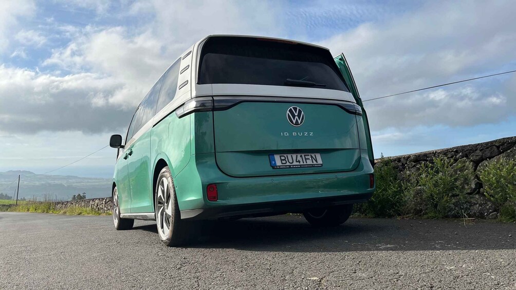 A green Volkswagen ID. Buzz van parked on a rural road with a stone wall and vegetation on the side. 3