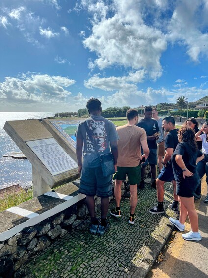 A group of people standing by a stone monument with an informational plaque, overlooking a body of water and a hilly landscape under a partly cloudy sky. 2