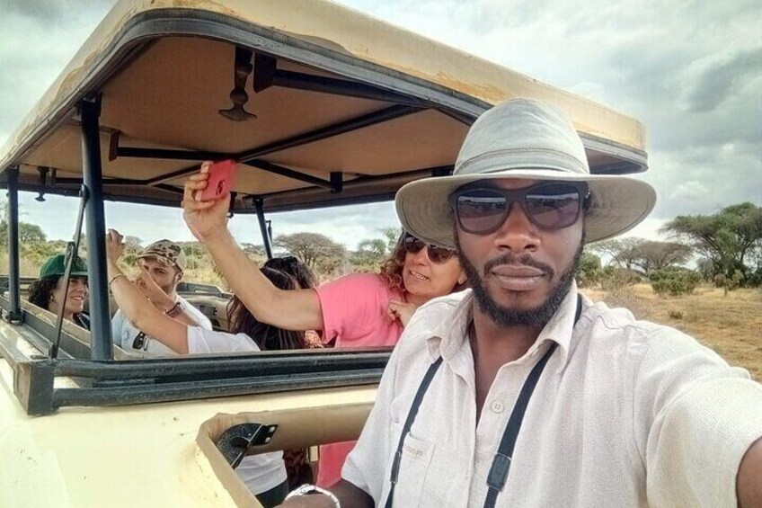 A safari vehicle with passengers, including a person in a white shirt and hat, and another in a pink shirt. 1