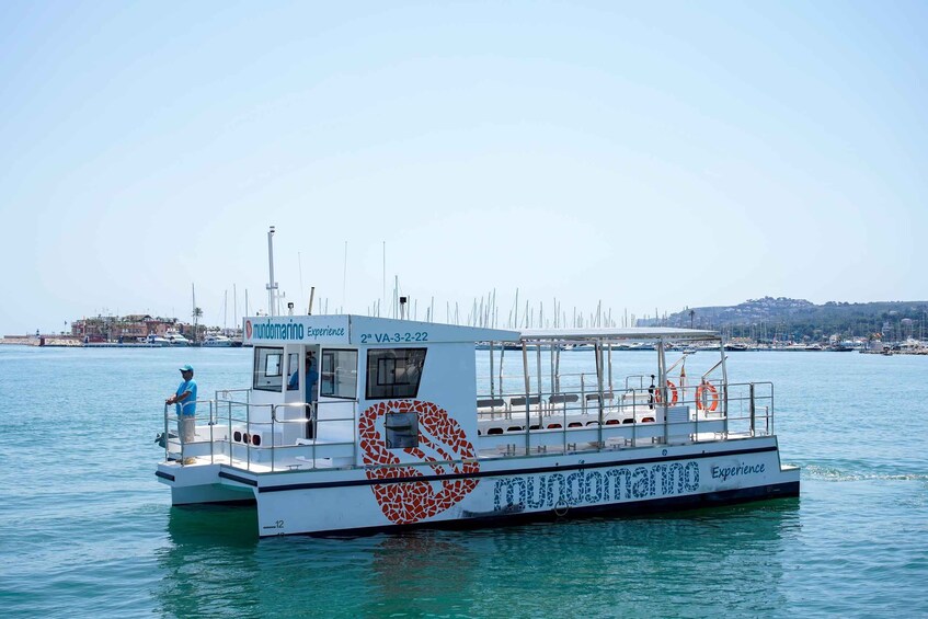 A boat with a heart-shaped design and the text 'mundomarina Experience' on its side, docked at a marina with other boats and a distant shoreline. 5