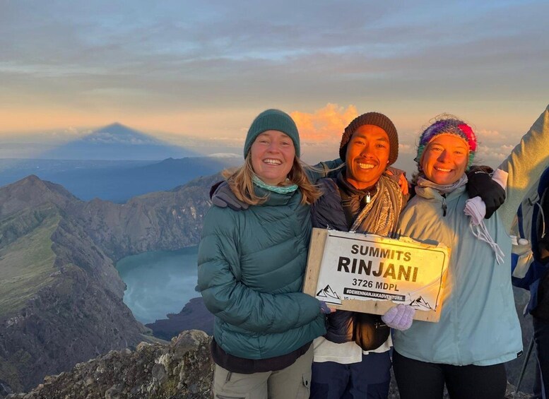 Three people standing on a mountain with a sign that reads "Summits Rinjani 3726 MDPL". 1