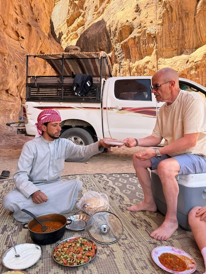 Two men sitting on a mat outdoors with food and a white pickup truck in the background. 4