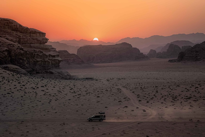 A desert landscape with a vehicle driving on a dirt road, surrounded by rocky formations and a sunset in the background. 3