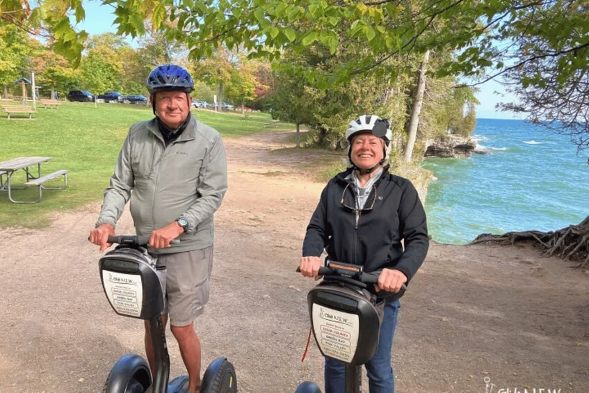 Two people on Segways near a body of water with trees and a picnic area in the background. 2