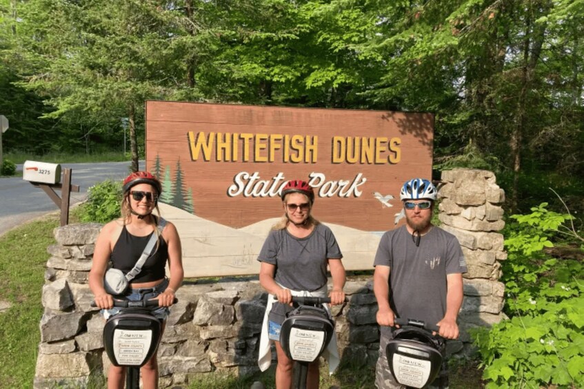 Three people on bicycles in front of a sign for Whitefish Dunes State Park. 3