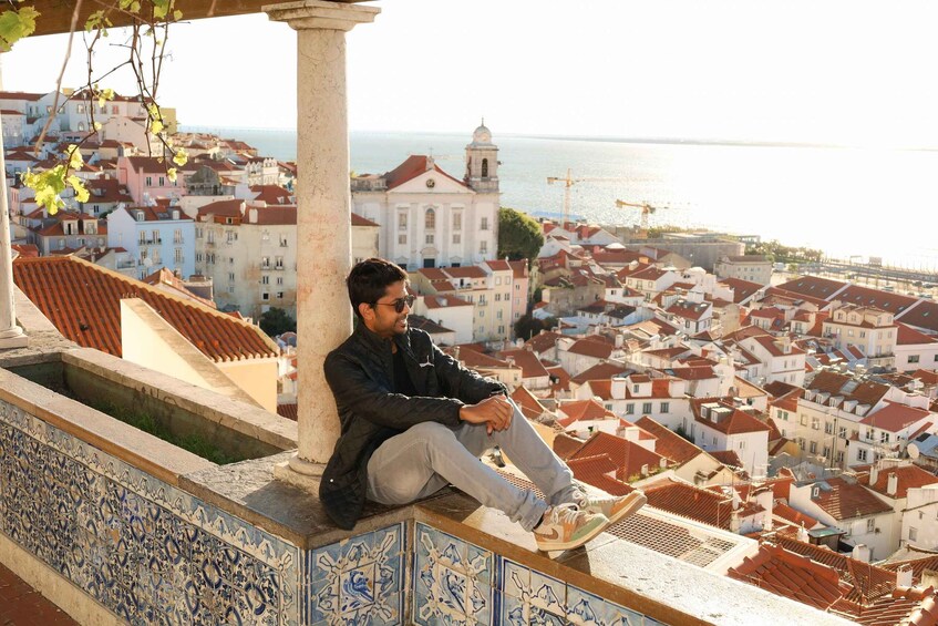 A man sitting on a tiled ledge overlooking a coastal town with red-roofed buildings and a body of water in the distance. 3