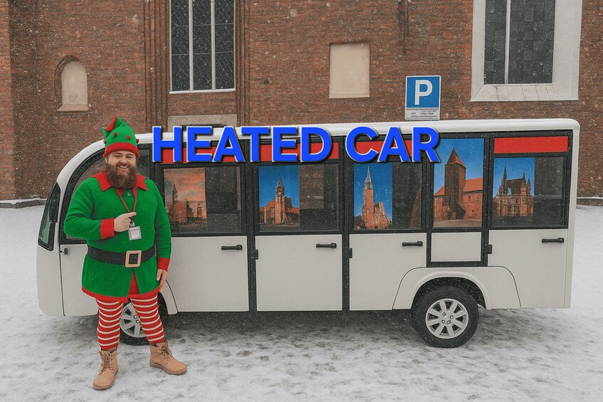 A person in a green elf costume standing next to a white bus with a sign that reads "HEATED CAR". 2