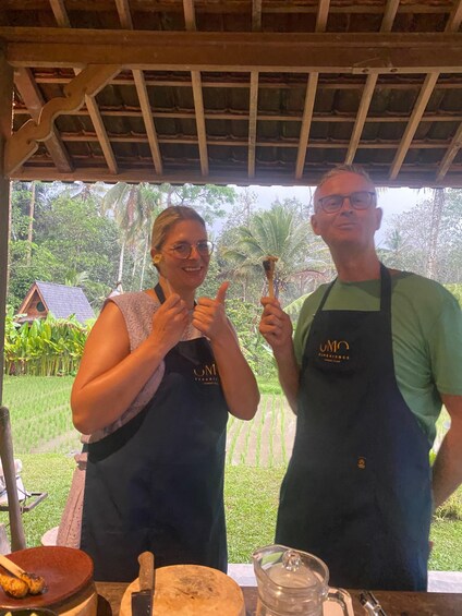 Two people wearing aprons with a logo, standing in a covered outdoor area with a table in front of them. 2