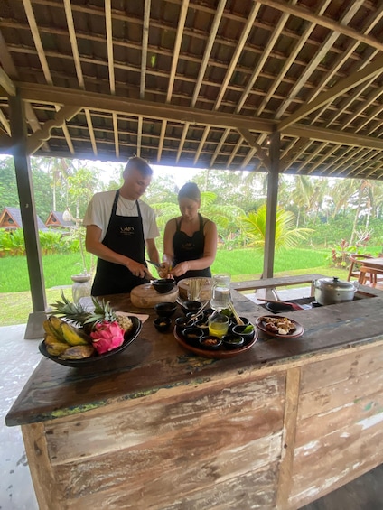 Two people preparing food at an outdoor wooden table with tropical plants in the background. 2