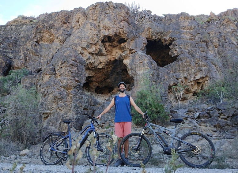 A person standing with two bicycles in front of a rocky landscape with a large cave-like opening. 2