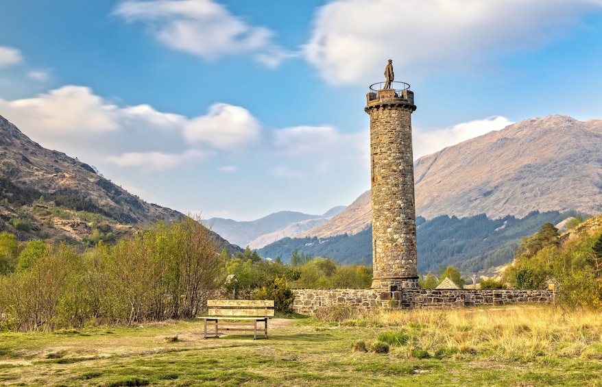 A stone tower with a statue on top, surrounded by mountains and greenery. 2