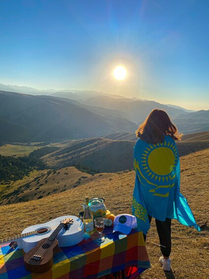 A person with a sun-patterned shawl overlooking a mountainous landscape with a guitar and a picnic setup. 1