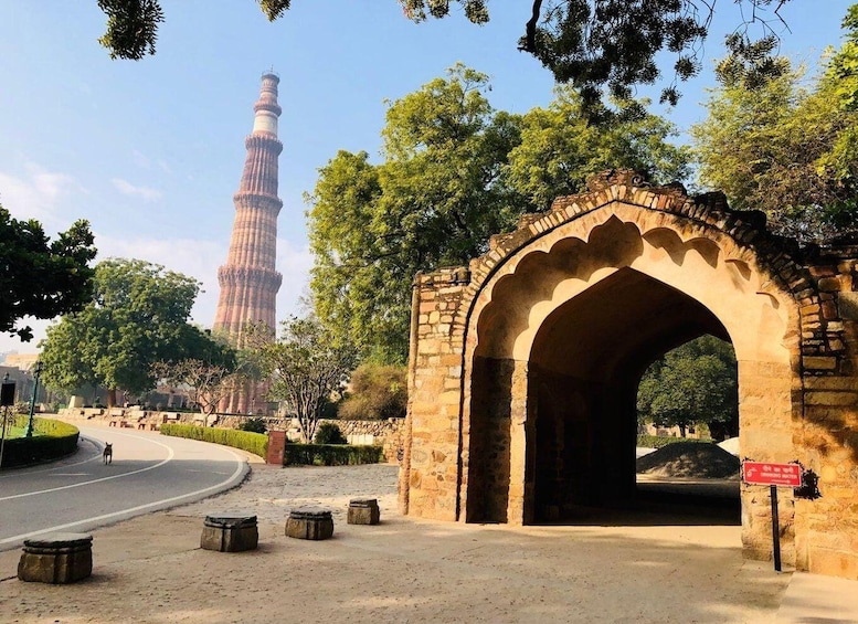 A historic archway with a sign, a road, and the Qutub Minar in the background. 1