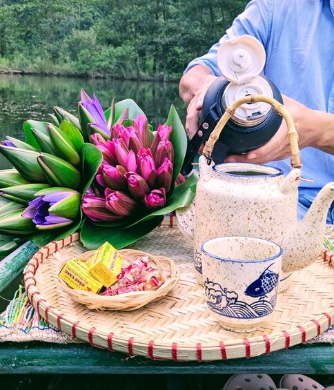 Eine Person gießt Tee aus einer traditionellen Teekanne in eine Keramiktasse, mit Lotusblumen und einer gewebten Matte auf einem Boot. 5