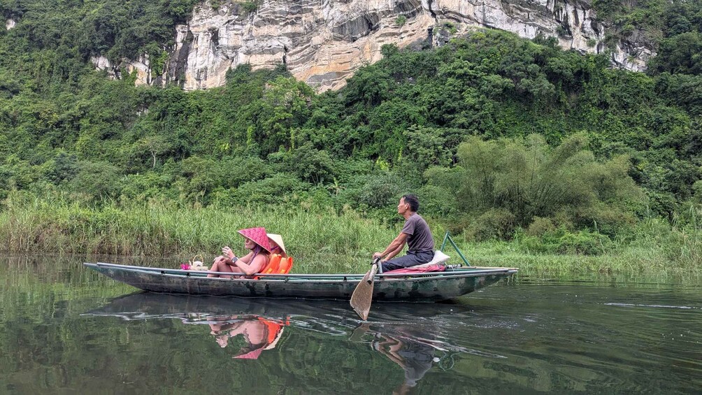 Ein Mann rudert ein Boot mit zwei Passagieren, umgeben von üppiger Vegetation und einer felsigen Klippe im Hintergrund. 2