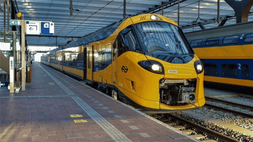 A yellow train at a station platform with overhead signs and tracks. 2