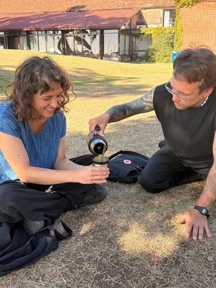 Two people outdoors, one pouring from a thermos, with a building and grass in the background. 1
