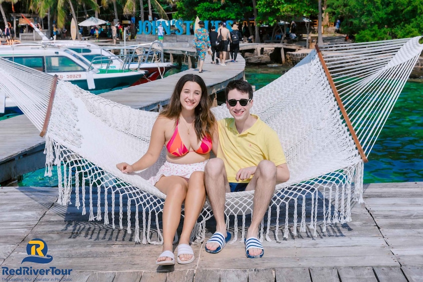 A couple sitting in a hammock on a wooden dock with a boat in the background. 1