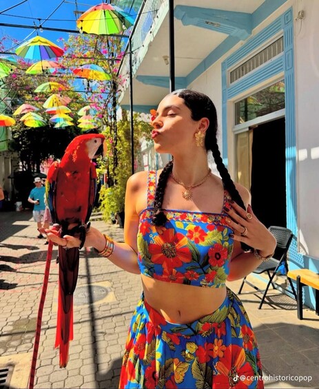 A person holding a red parrot in an outdoor setting with colorful umbrellas and a blue building in the background. 1