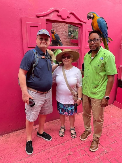 Three people standing in front of a pink building with a parrot perched on a window ledge. 3