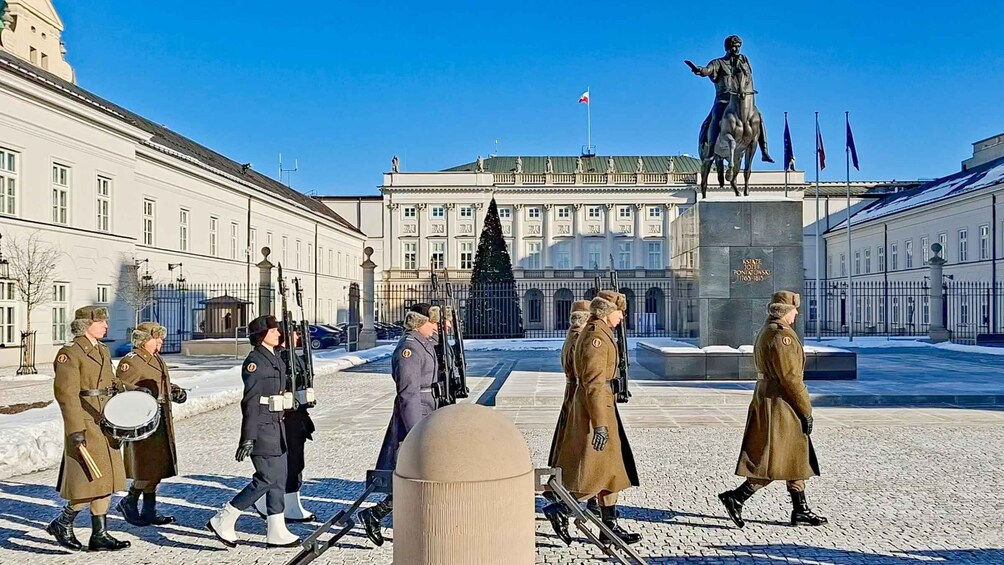 A group of uniformed individuals in a snowy courtyard with a statue and a building in the background. 1