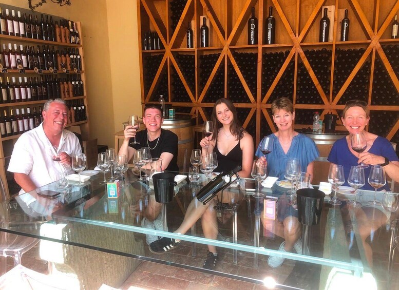 A group of people seated at a bar with wine glasses, bottles, and a wine rack in the background. 2
