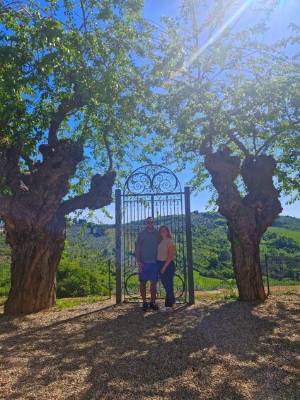 Two people standing under a wrought iron gate with a vineyard in the background. 4