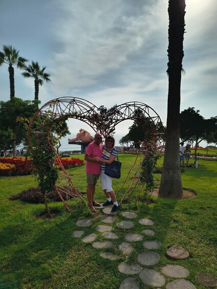 A couple standing under a heart-shaped structure in a park with palm trees and flower beds. 5