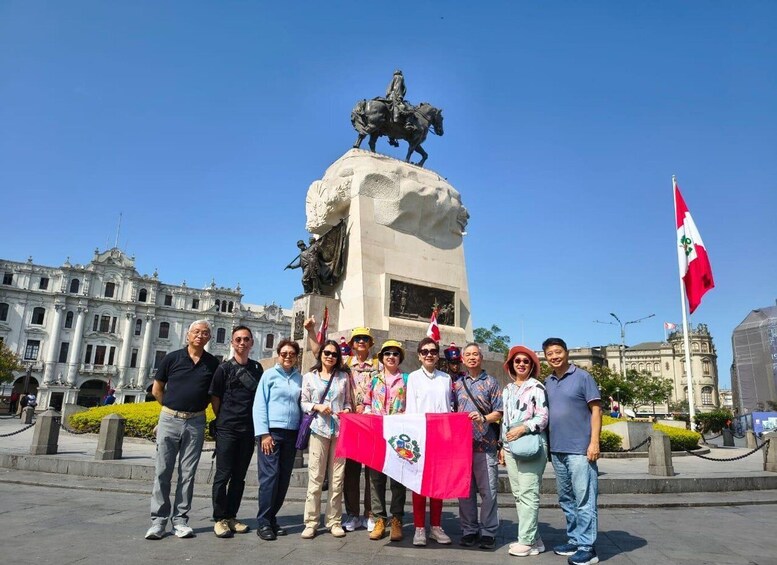 A group of people standing in front of a monument with a statue of a figure on horseback, flags, and a historic building in the background. 1