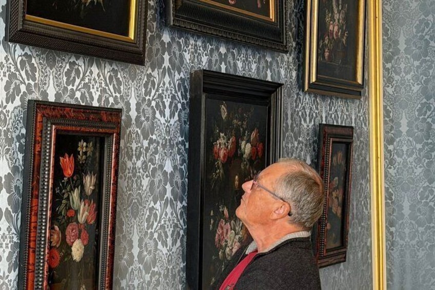 A man observing framed floral paintings on a wall with patterned wallpaper. 4