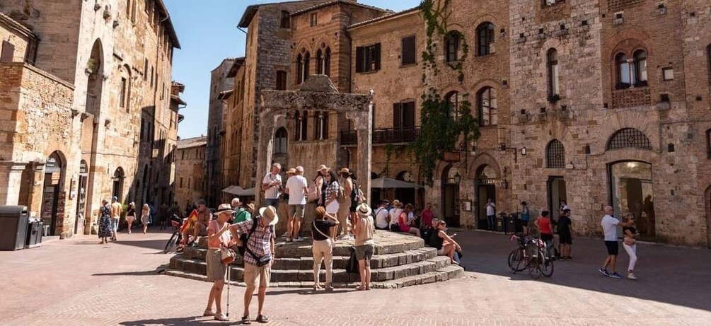 A historic square with stone buildings, a central fountain, and people walking and sitting. 1