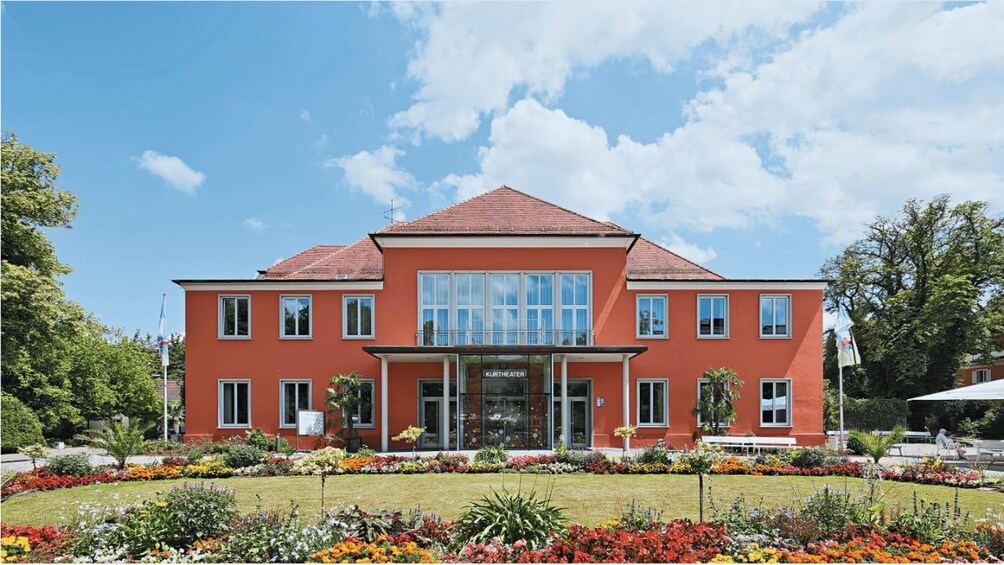 A red building with a sign reading 'KURTHEATER' in front of a well-maintained garden with colorful flowers. 2