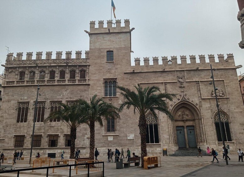 A historic building with a tower and multiple windows, palm trees in the foreground, and people walking around. 5