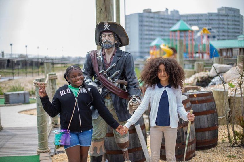 Eine Piratenstatue, ein Kind mit Piraten-Kapuze und ein weiteres Kind mit Spielschwert in einem Park mit Spielplatz im Hintergrund. 2