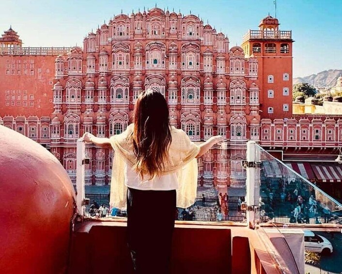 A person standing with arms outstretched, overlooking a large, ornate building with multiple levels and intricate architectural details. 3