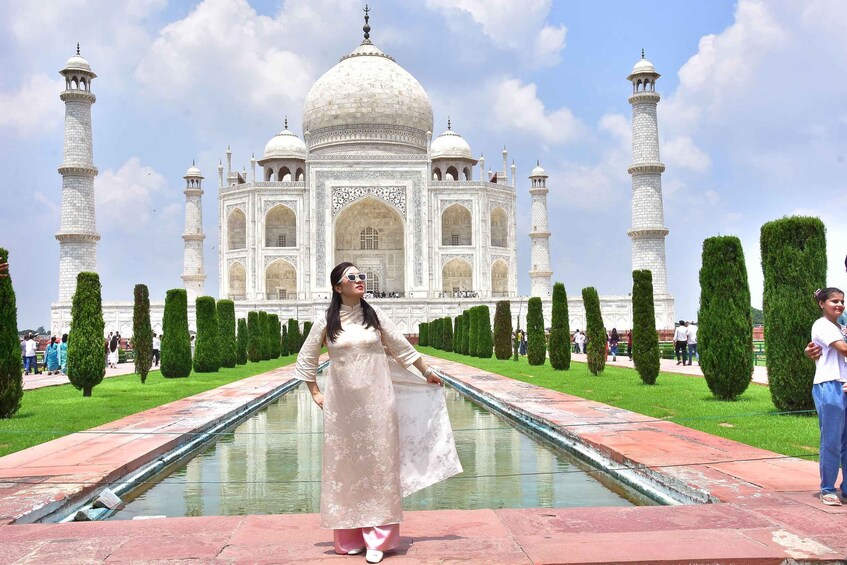 A person in a dress standing in front of the Taj Mahal with a pool and neatly trimmed trees. 4