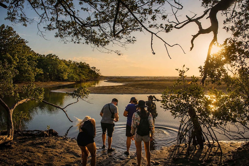 A group of people standing by a calm river at sunset, surrounded by trees and a sandy shore. 3