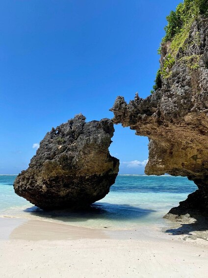 A rock formation arching over the ocean with clear blue water and a sandy beach. 3