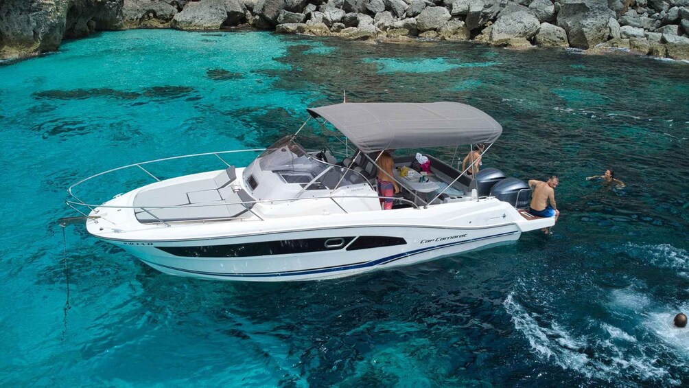A white motorboat with a grey canopy, partially submerged in clear turquoise water near a rocky shore. 3