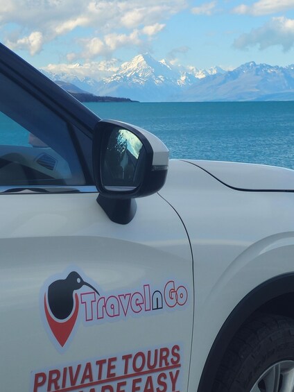 Valkoinen auto, jonka kylkeen on maalattu tekstit »TravelInGo» ja »PRIVATE TOURS EASY», on pysäköitynä vesistön ääreen, ja taustalla näkyy vuoria. 2