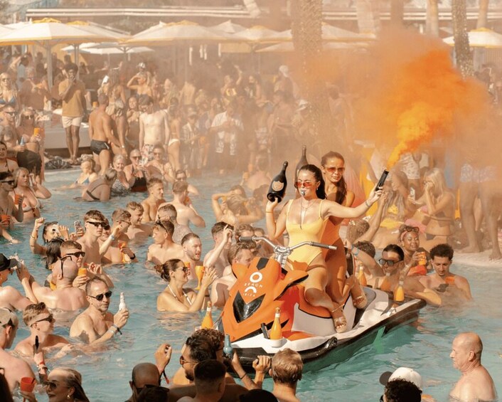 A crowded pool with people in the water and a boat with two people riding it. 2