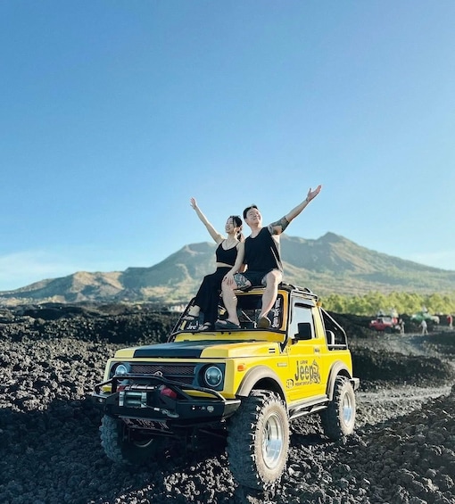 Two people on a yellow off-road vehicle with raised arms, driving on a rocky terrain with a mountain in the background. 2
