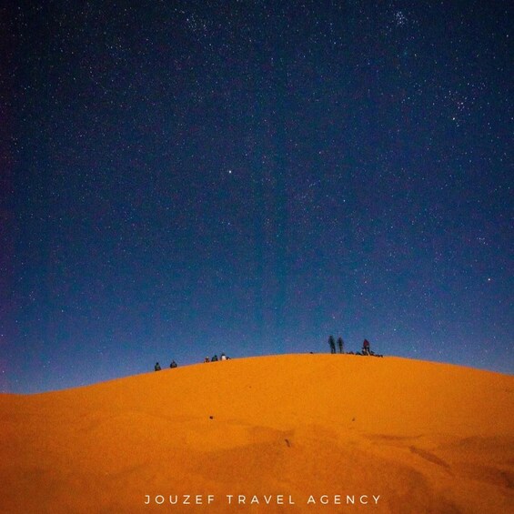 A desert landscape with a starry night sky and a group of people on a sand dune. 2