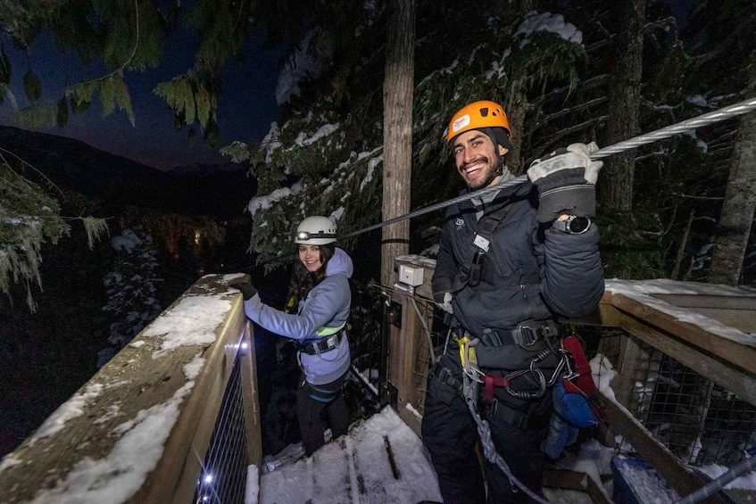 Two people in helmets and harnesses on a snowy bridge with trees in the background. 3