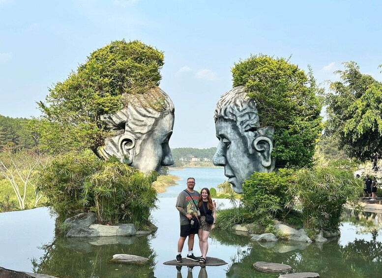 A couple standing in front of a sculpture with two faces, surrounded by water and greenery. 1