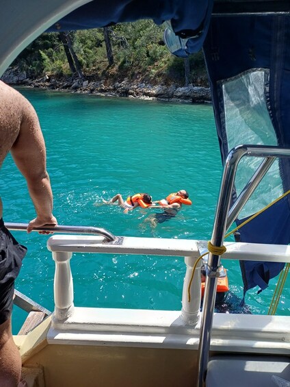A person swimming in turquoise water, wearing an orange life jacket, with a boat and rocky shoreline in the background. 5